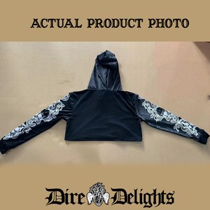 May include: Black cropped hoodie with a white graphic print of a skull and snakes on the sleeves. The hoodie has a drawstring hood and a relaxed fit.