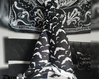 May include: Black and white leggings with a bat print. The leggings are worn by a person sitting on a bed with a black and white tapestry behind them.