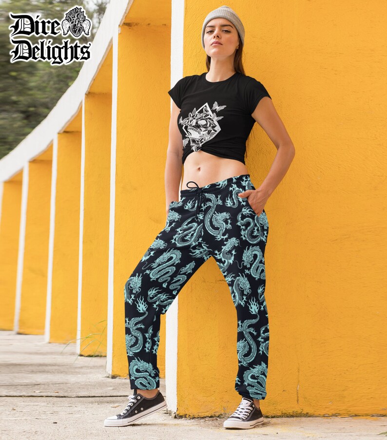 May include: A black t-shirt with a white graphic and dragon print joggers. The joggers are black with a teal dragon pattern. The model wears a grey beanie and black trainers. The logo "Dire Delights" is in the top left corner.