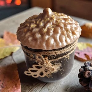 May include: An acorn-shaped candle with a brown base and a textured, light brown top sprinkled with gold glitter. The candle is wrapped with twine and a decorative cutout. Autumn leaves and a pinecone are scattered around it.