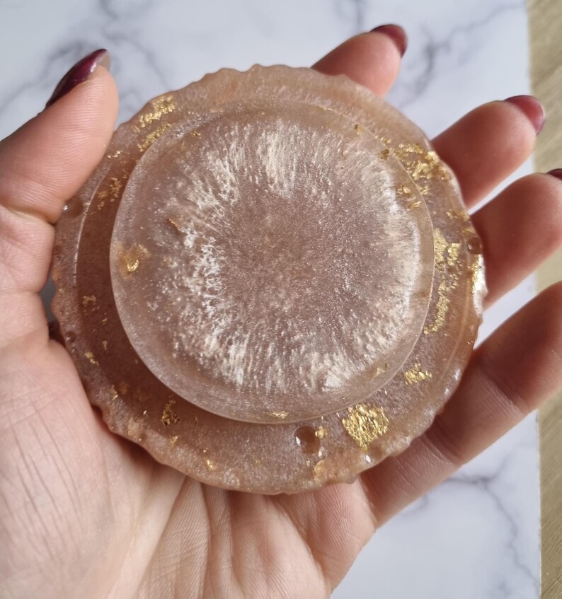 May include: A set of three resin coasters in shades of pink and white, with gold leaf accents. The coasters have a glossy finish and a circular shape, perfect for protecting surfaces from drink rings.