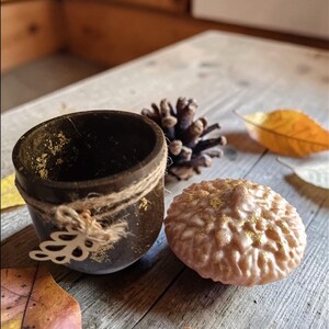 May include: A dark brown, small, round container with gold flecks and twine detailing, next to a light brown, textured, round object with gold accents. A pine cone and autumn leaves are also present, all on a wooden surface.