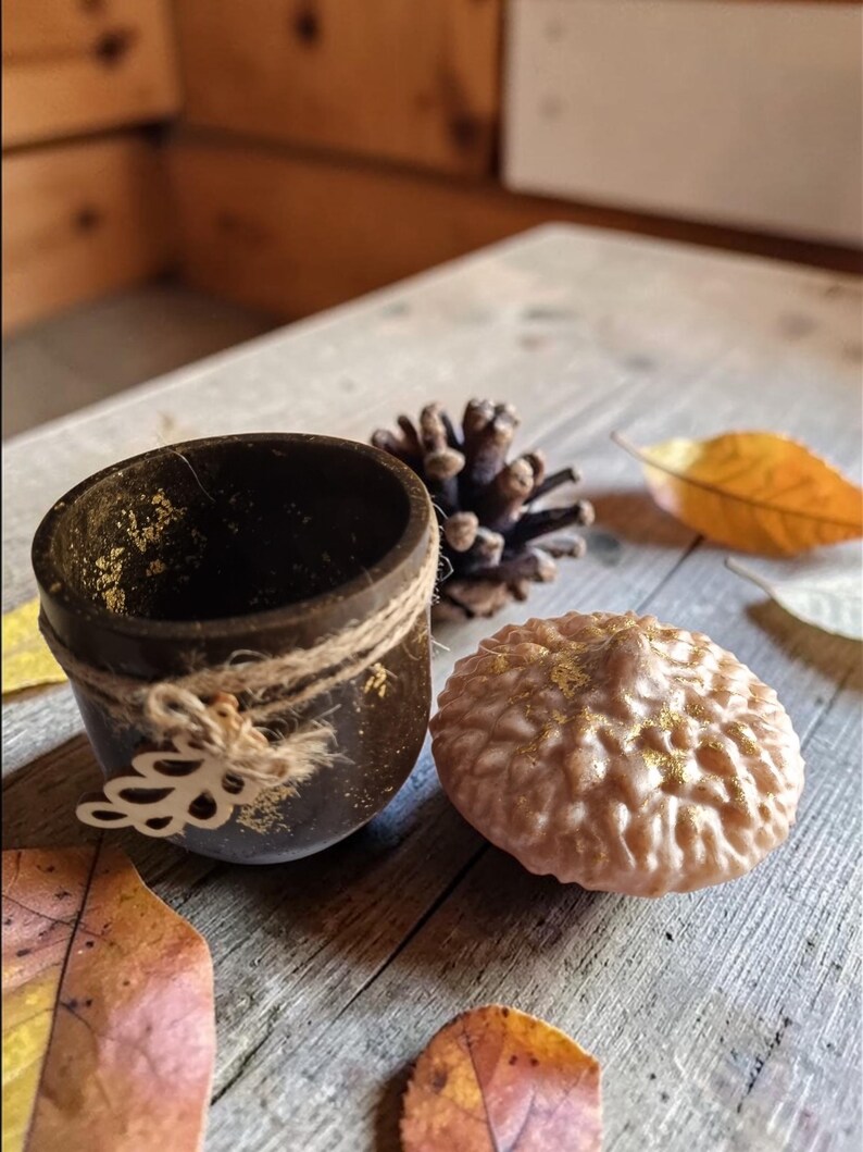May include: A dark brown, small, round container with gold flecks and twine detailing, next to a light brown, textured, round object with gold accents. A pine cone and autumn leaves are also present, all on a wooden surface.