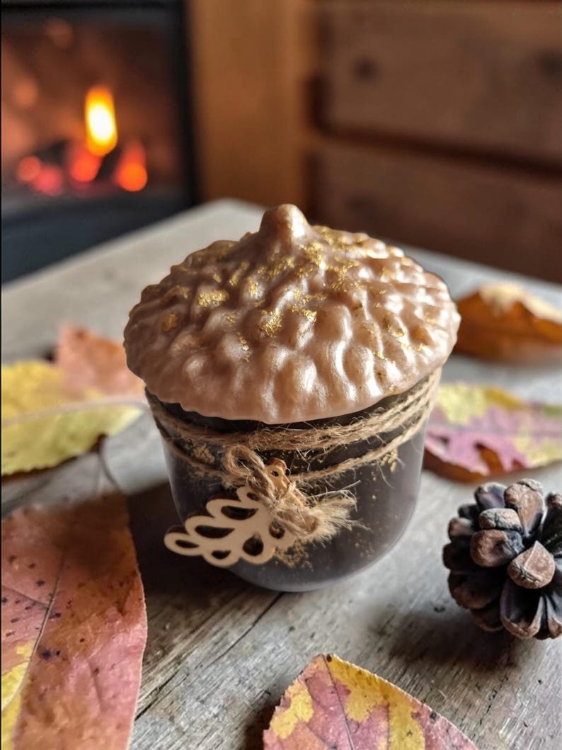 May include: An acorn-shaped candle with a brown base and a textured, light brown top sprinkled with gold glitter. The candle is wrapped with twine and a decorative cutout. Autumn leaves and a pinecone are scattered around it.
