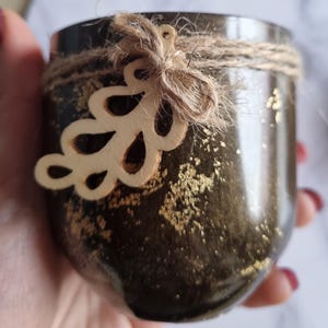 May include: A dark brown jar with gold flecks, wrapped with twine and a wooden leaf embellishment. The jar has a rustic, handmade appearance, suitable for candles or small plants. The design is simple and elegant.