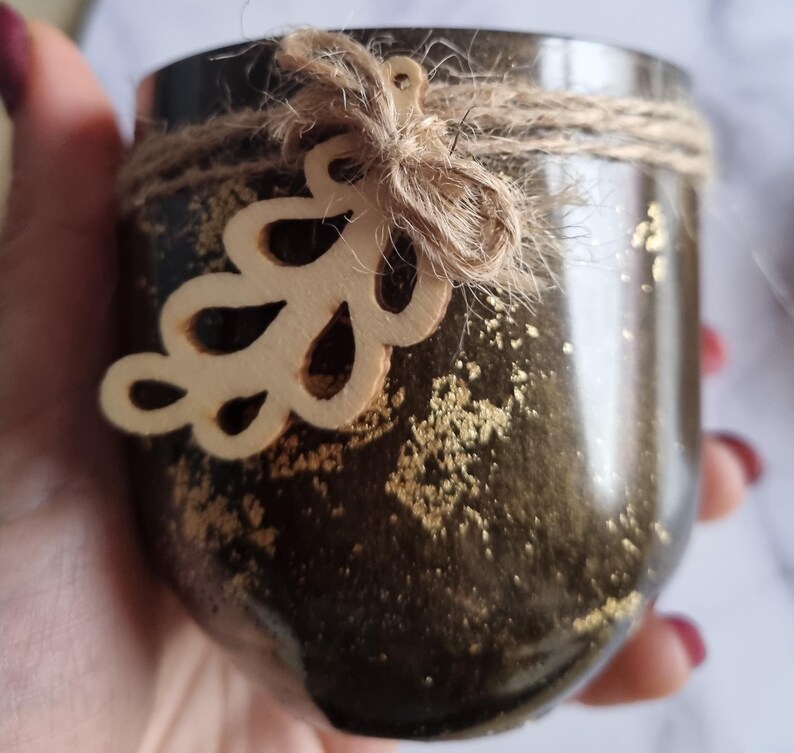 May include: A dark brown jar with gold flecks, wrapped with twine and a wooden leaf embellishment. The jar has a rustic, handmade appearance, suitable for candles or small plants. The design is simple and elegant.