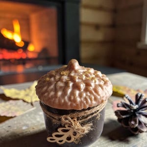 May include: A brown acorn-shaped candle with a textured top and a dark brown base, tied with twine and a decorative leaf. The candle sits on a wooden surface with a pine cone and autumn leaves. A fireplace with a warm glow is in the background.