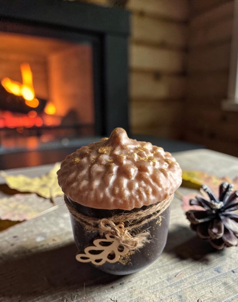 May include: A brown acorn-shaped candle with a textured top and a dark brown base, tied with twine and a decorative leaf. The candle sits on a wooden surface with a pine cone and autumn leaves. A fireplace with a warm glow is in the background.