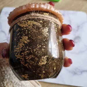 May include: A small, dark brown jar with gold leaf accents and a natural twine tie. The jar has a light brown, textured rim and is held in a person's hand. The jar appears to be a decorative item or container.