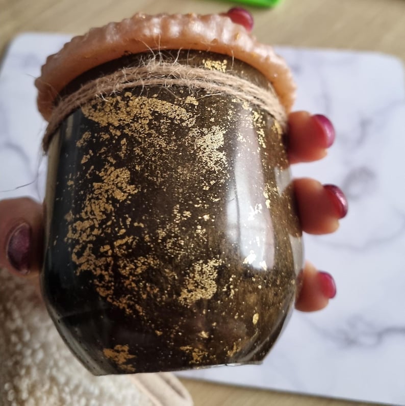 May include: A small, dark brown jar with gold leaf accents and a natural twine tie. The jar has a light brown, textured rim and is held in a person's hand. The jar appears to be a decorative item or container.