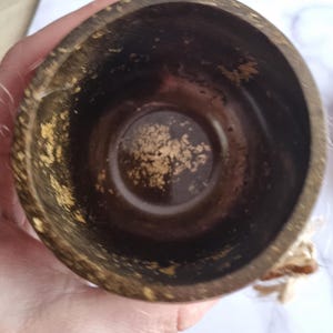 May include: A dark brown, handcrafted wooden cup with gold accents. The interior of the cup is a deep brown, and the rim and exterior feature flecks of gold. The cup is held in a hand, with a blurred background.
