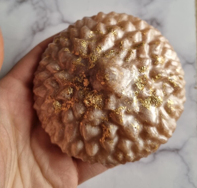 May include: A round, textured soap bar with a light brown color and gold flecks. The soap has a bumpy surface, resembling a pinecone. The soap is held in a hand.