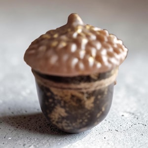 May include: A detailed close-up of a decorative acorn. The acorn's cap is light brown with gold accents, while the body is dark brown with gold speckles. The acorn is set against a light gray background.