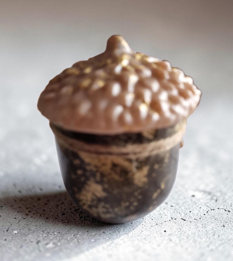 May include: A detailed close-up of a decorative acorn. The acorn's cap is light brown with gold accents, while the body is dark brown with gold speckles. The acorn is set against a light gray background.