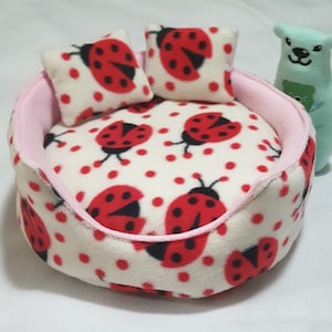 May include: A pink and white round pet bed with a ladybug print. The bed has a pink rim and two matching ladybug print cushions.
