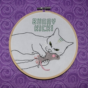 以下が含まれることがあります： 白い猫が灰色のネズミに飛びかかる刺繍のフープアート。猫の上に緑色の文字で「Bunny Kick!」と刺繍されています。