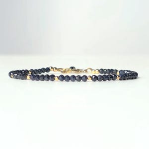Könnte beinhalten: Ein zartes Armband mit kleinen, facettierten, dunkelblauen Perlen, die mit goldfarbenen Perlen durchsetzt sind. Das Armband ist auf einem dünnen, flexiblen Band aufgezogen und hat einen goldfarbenen Verschluss.