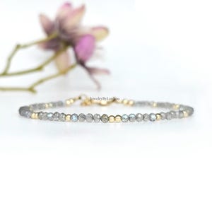 Op de afbeelding: Een fijne armband met kleine, facetgeslepen grijze kralen afgewisseld met goudkleurige kralen. De armband staat tegen een witte achtergrond met een vage bloem. Het is een sieraad.
