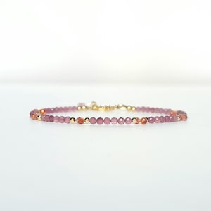 Op de afbeelding: Een delicate kralenarmband met kleine, gefacetteerde roze en oranje edelsteenkralen afgewisseld met piepkleine, gepolijste goudkleurige metalen bolletjes. De armband heeft een goudkleurige metalen sluiting en een label met de tekst JewelryByLouLou.