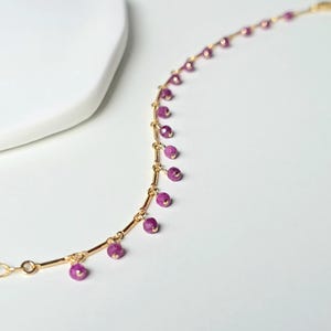 Può includere: Un delicato braccialetto dorato con piccole perline rotonde color magenta. Le perline sono uniformemente distanziate e pendono dalla catena d'oro. Il braccialetto è presentato su una superficie bianca, evidenziando il suo design elegante.