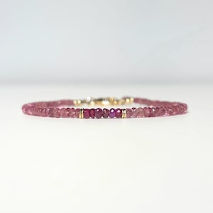 Può includere: Un braccialetto delicato con piccole perline di tormalina rosa sfaccettate disposte in una fascia circolare. Il centro è accentuato da un gruppo di perline color magenta più intenso, fiancheggiato da due piccoli distanziatori in metallo dorato e una chiusura.