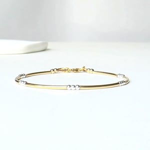 Könnte beinhalten: Ein gold- und silberfarbenes Armband mit filigranem Design. Das Armband hat ein dünnes, goldenes Band mit silbernen Akzenten und einem Verschluss. Der Schmuck wird auf einer weißen Oberfläche präsentiert.