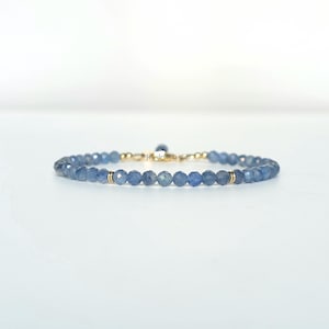 Könnte beinhalten: Ein zartes Armband mit kleinen, facettierten blauen Edelsteinperlen, die in einem Kreis angeordnet sind. Das Design umfasst winzige goldfarbene Metallabstandshalter zwischen den blauen Perlen und einen goldfarbenen Karabinerverschluss.
