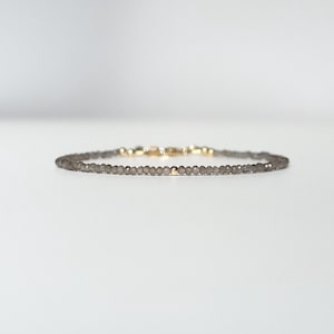 May include: A delicate bracelet composed of small, faceted grey beads arranged in a single strand. The bracelet features a single gold-toned bead at the centre and a gold-toned metal clasp mechanism visible at the back of the circular arrangement.