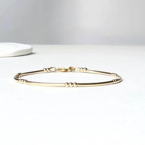 Könnte beinhalten: Ein zartes Goldarmband mit schlichtem Design. Das Armband hat einen schlanken, gebogenen Stab mit kleinen, dekorativen Perlen und einem Verschluss. Der Schmuck ist vor einem weißen Hintergrund platziert.