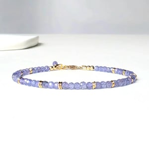 Könnte beinhalten: Ein zartes Armband mit kleinen, facettierten, hellvioletten Perlen. Das Armband ist mit goldfarbenen Metallabstandshaltern und einem Verschluss versehen. Der Schmuck wird auf einer weißen Oberfläche präsentiert.