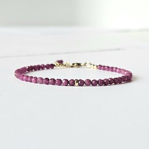 Könnte beinhalten: Ein zartes Armband mit kleinen, facettierten, tiefrosa Edelsteinen. Das Armband hat goldfarbene Akzente und einen Verschluss. Der Schmuck wird auf einer weißen Oberfläche präsentiert.