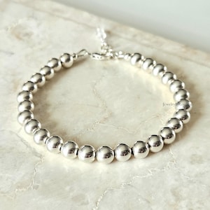 Könnte beinhalten: Ein silbernes Perlenarmband mit einer zarten Kette und einem Verschluss. Das Armband besteht aus mehreren runden, glänzenden Perlen, die aneinandergereiht sind. Der Schmuck wird auf einer hellen Oberfläche präsentiert.