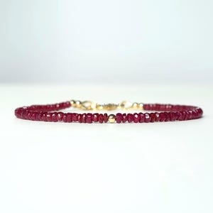 Könnte beinhalten: Ein zartes Armband mit kleinen, facettierten, tiefroten Rubinperlen. Das Armband ist an einer Goldkette befestigt und hat einen Goldverschluss. Der Schmuck wird auf einer weißen Oberfläche präsentiert.