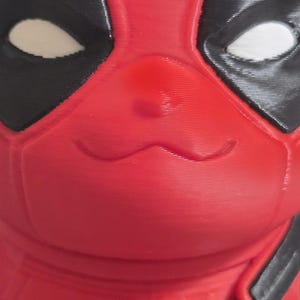 May include: A red and black figurine featuring a character with a cat-like face, wearing a mask with black eye patches and white eye cutouts. The figure has a smooth, molded surface with a diagonal black strap across the chest area.