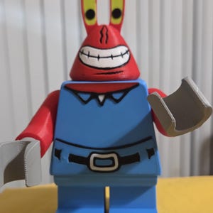 May include: A plastic toy figure resembling a crab character with a red head, tall eye stalks, and a wide grin. The figure wears a blue shirt with a black belt and collar detail, red arms, and grey claw-shaped hands. The design is blocky and articulated.