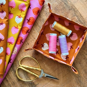 May include: A fabric sewing project with a small tray lined with orange fabric with a strawberry pattern. The tray contains three spools of thread: gold, pink, and purple. A pair of gold scissors is on the wooden surface next to the tray.