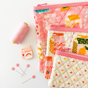 May include: Pink quilted zipper pouches with milk carton designs, a spool of pink thread, a milk carton pin, and pink-headed pins. The pouches have pink zippers and are in various sizes, with heart and fruit patterns.