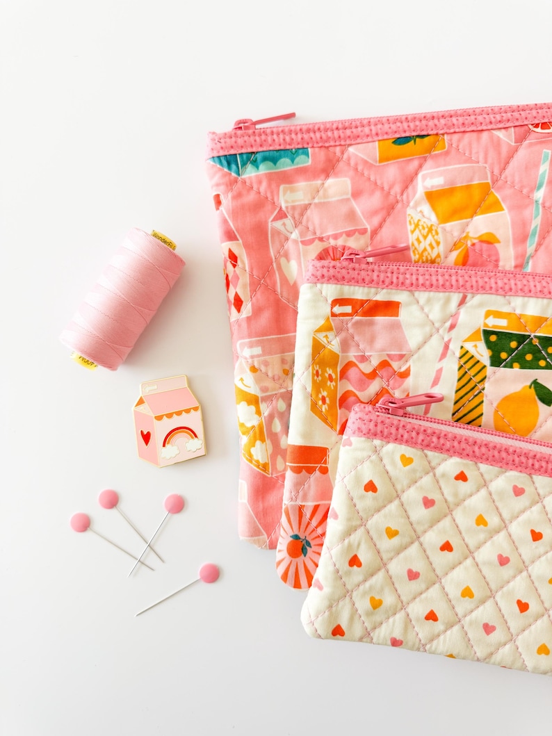 May include: Pink quilted zipper pouches with milk carton designs, a spool of pink thread, a milk carton pin, and pink-headed pins. The pouches have pink zippers and are in various sizes, with heart and fruit patterns.