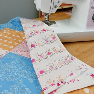 May include: A white sewing machine with the word "brother" printed on the side is sewing a quilt. The quilt is made of different fabrics with a variety of patterns, including a blue fabric with a geometric pattern, a pink fabric with a floral pattern, and a white fabric with a pattern of mice wearing dresses.