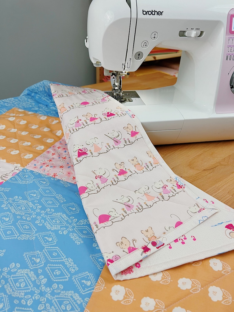 May include: A white sewing machine with the word "brother" printed on the side is sewing a quilt. The quilt is made of different fabrics with a variety of patterns, including a blue fabric with a geometric pattern, a pink fabric with a floral pattern, and a white fabric with a pattern of mice wearing dresses.