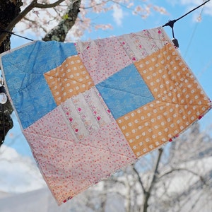 May include: A patchwork quilt with a variety of floral and geometric patterns in pink, blue, and yellow. The quilt is hanging on a clothesline outdoors.