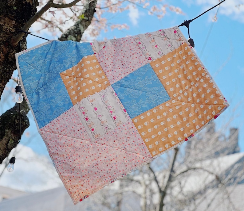 May include: A patchwork quilt with a variety of floral and geometric patterns in pink, blue, and yellow. The quilt is hanging on a clothesline outdoors.