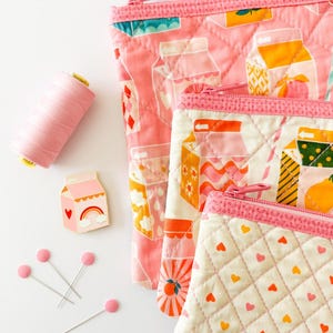 May include: A collection of pink quilted pouches with zipper closures, featuring milk carton and heart patterns. A spool of pink thread, a milk carton pin, and pink-headed pins are also present. The Stacey Lee Creative logo is in the upper left corner.