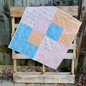 May include: A patchwork quilt with a blue, pink, white, and orange pattern. The quilt is folded and leaning against a wooden pallet.