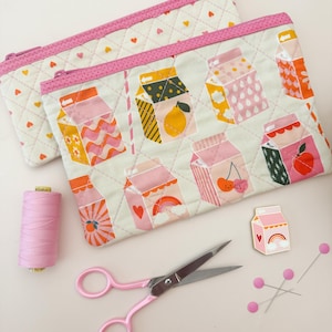 May include: Two quilted pencil cases with a milk carton pattern in various colors, including pink, orange, and yellow. The cases have pink zippers and are next to pink scissors, thread, pins, and a milk carton pin.