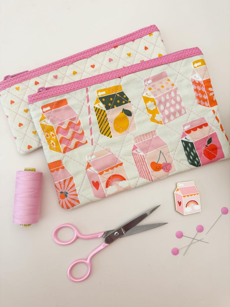 May include: Two quilted pencil cases with a milk carton pattern in various colors, including pink, orange, and yellow. The cases have pink zippers and are next to pink scissors, thread, pins, and a milk carton pin.