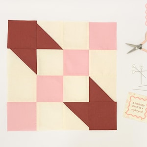 May include: A patchwork quilt block in a checkerboard pattern with burgundy, light pink, and cream-colored squares. A pair of small scissors, pins, and a card that reads "A finished quilt is a perfect gift" are also present.