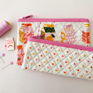 May include: Two quilted fabric pouches with pink zippers and trim. One pouch features a pattern of milk carton illustrations, and the other has a heart-shaped pattern. A spool of pink thread, pink pins, and a milk carton pin are also in the image.
