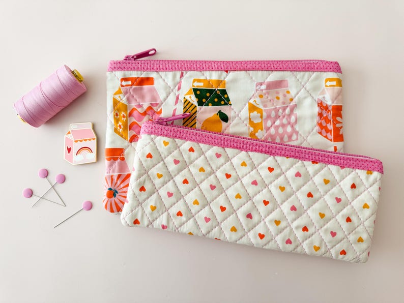 May include: Two quilted fabric pouches with pink zippers and trim. One pouch features a pattern of milk carton illustrations, and the other has a heart-shaped pattern. A spool of pink thread, pink pins, and a milk carton pin are also in the image.