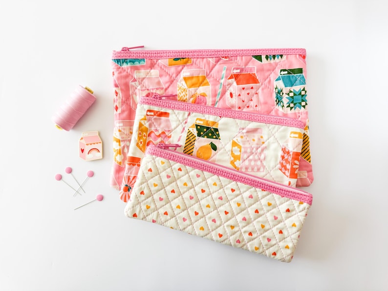 May include: Three quilted zipper pouches in varying sizes, featuring pink and white fabrics with colorful milk carton and heart patterns. A spool of pink thread, pins, and a milk carton pin are also present.